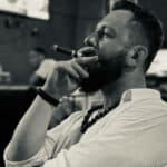 Man smoking cigar, wearing necklace and white shirt.