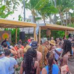 Crowded beach party with DJ and tropical scenery.